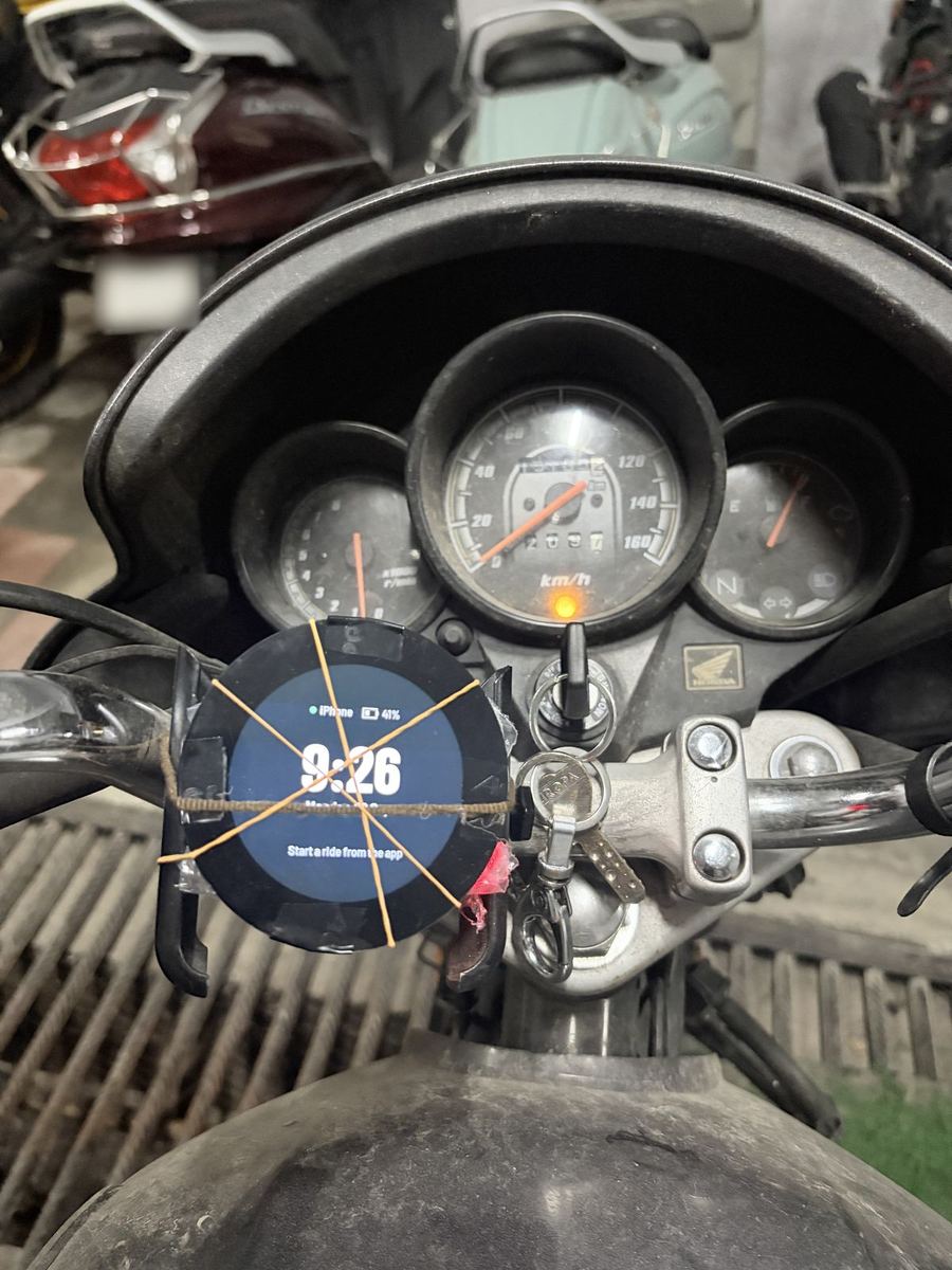 Round display tied to a motorcycle handlebar with rubber bands, next to the speedometer